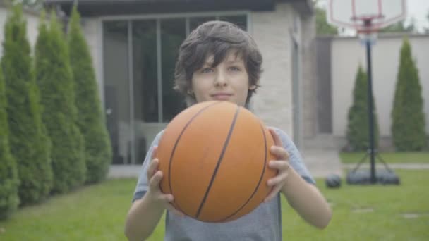 Porträtt av leende självsäker vit pojke sträcker basket framför kameran. Glada lilla barn poserar med sportutrustning utomhus. Söt brunett unge angelägen om sport. — Stockvideo