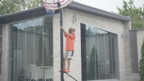 Grande tiro de menino engraçado escalando no basquete ao ar livre. Retrato de um miúdo caucasiano a divertir-se no quintal. Criança descansando aos fins de semana. Infância e lazer. — Vídeo de Stock