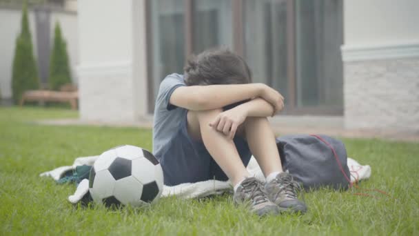 Depressiver kleiner brünetter Junge, der mit Fußball auf dem Rasen sitzt. Porträt eines gestressten kaukasischen Kindes, das den Kopf auf die Knie legt und das Gesicht hebt. Süß schoolboy having problems. — Stockvideo