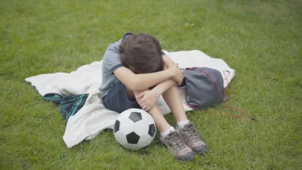 Vista de ángulo superior de niño caucásico frustrado sentado con la cabeza sobre las rodillas, empujando la pelota de fútbol, y dejando. Retrato de niño deprimido en césped verde al aire libre. Niño fracasado. — Vídeos de Stock