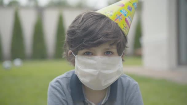 Κοντινό πλάνο του θλιμμένου Καυκάσιου μελαχρινού αγοριού με μάσκα προσώπου και καπέλο για πάρτι κοιτάζοντας την κάμερα. Πορτρέτο δυστυχισμένου μικρού παιδιού που γιορτάζει μόνος του τα γενέθλιά του στην πανδημική καραντίνα Covid-19. — Αρχείο Βίντεο