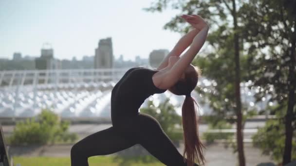 カメラは、都会を背景に日の出に逆曲げ若いスリムな白人女性の動きに従います。午前中の魅力的な柔軟な体操の側面図. — ストック動画