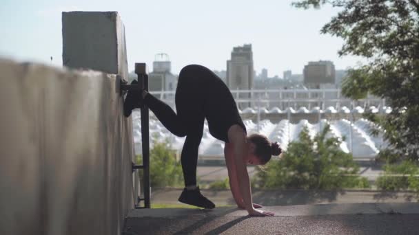 Jonge vrouwelijke turnster of yogi doen handstand met benen gespleten buiten. Slanke flexibele blanke vrouw training op de achtergrond van de stad bij zonsopgang. — Stockvideo