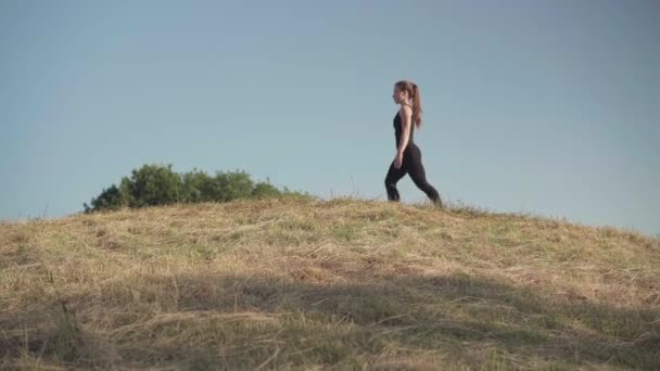 Amplo tiro de ginasta feminina caucasiana andando ao longo da colina e pulando para cima levantando a perna. Retrato de vista lateral de mulher incrível flexível treinando ao ar livre no fundo do belo céu azul de verão. — Vídeo de Stock
