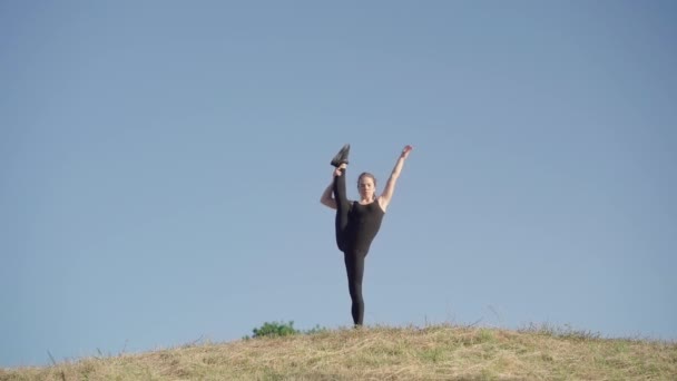 Portrait of extremely flexible young woman raising leg up. Wide shot of Caucasian sportswoman warming up outdoors standing on hill. Elasticity and plasticity of human body. — Stock Video