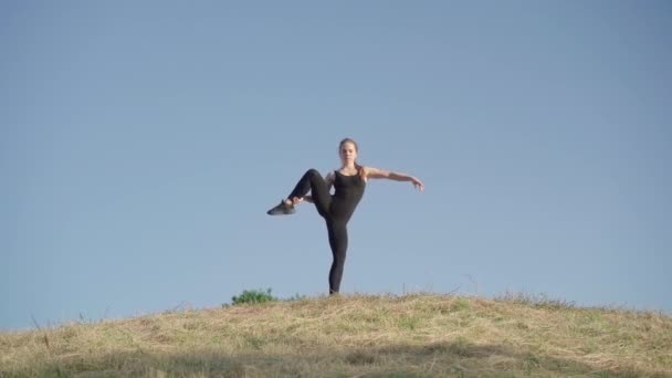 자신감 있고 유연 한 젊은 여성이 맑은 푸른 하늘 배경에 서 있습니다. 운동 선수의 모습을 그린 카프카스 체조 선수나요기가 야외의 언덕에서 몸을 따뜻하게 해 주는 모습. — 비디오
