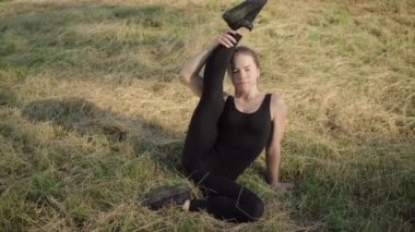 Gülümseyen beyaz kadın bacağını kaldırıp kameraya bakıyor. Esnek kadın jimnastikçinin portresi parkta çimlerin üzerinde egzersiz yapıyor. Sportif yaşam tarzı.