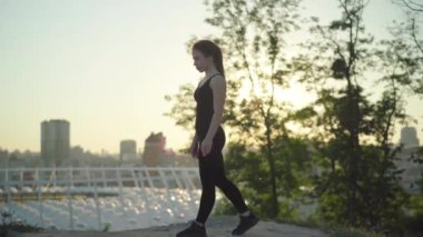 Gün batımında yürüyen ve zıplayan kendine güvenen beyaz kadın jimnastikçi. Arka planda şehir şehriyle gün ışığında antrenman yapan zayıf genç bir kadının yan görüntüsü.