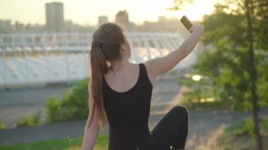 Gün batımında Park Hill 'de oturan ve selfie çeken beyaz bir kadının arka görüntüsü. Kaygısız sıska kadın arka planda kentsel şehirle birlikte yaz akşamının tadını çıkarıyor..