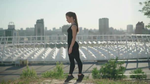 Sidovy av självsäker gymnast hoppar upp på bakgrunden av urban stad skyskrapor. Bred bild av vacker smal vit kvinna i svart uniform utövar utomhus. — Stockvideo