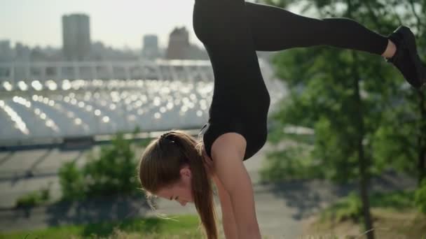 A câmera se move para cima e para baixo ao longo do corpo magro flexível da jovem mulher caucasiana de pé em mãos com as pernas para cima. Forte confiante ginasta feminina alongamento em raios de sol ao ar livre com a cidade no fundo. — Vídeo de Stock