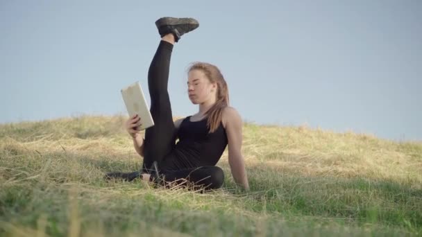 젊고 날씬 한 여자가 다리를 들고 언덕 위에 앉아 태블릿 셀카 카메라로 이야기하고 있습니다. 유연하고 놀라운 코카서스 체조 선수 온라인 무선 채팅을 사용하여 아침 야외에서 훈련으로. — 비디오