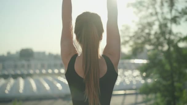 Pohled zezadu na štíhlou bělošskou brunetku s culíkem, stojící ve slunečních paprscích se zdviženýma rukama. Mladá sportovkyně si užívá ranní trénink venku s městem v pozadí. — Stock video