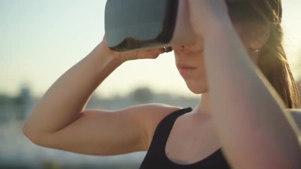 Close-up van een jonge brunette die VR-brillen in zonnestralen afdoet. Side view portret van zelfverzekerde blanke vrouwelijke gamer met behulp van augmented reality buiten bij zonsondergang. — Stockvideo