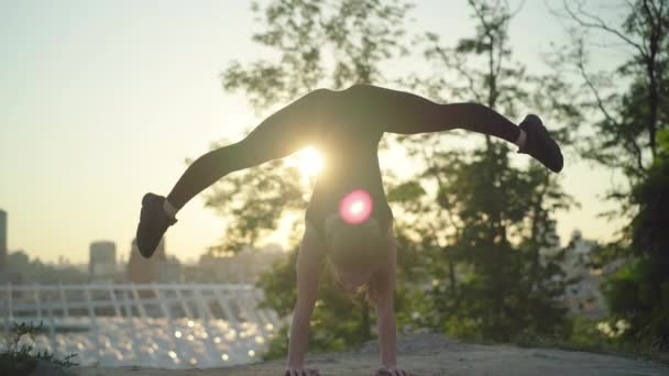 Breed schot handstand van slanke jonge turner in zonnestralen met wazig stad op de achtergrond. benen gespleten pose van slanke flexibele blanke vrouw bij zonsondergang buiten. — Stockvideo