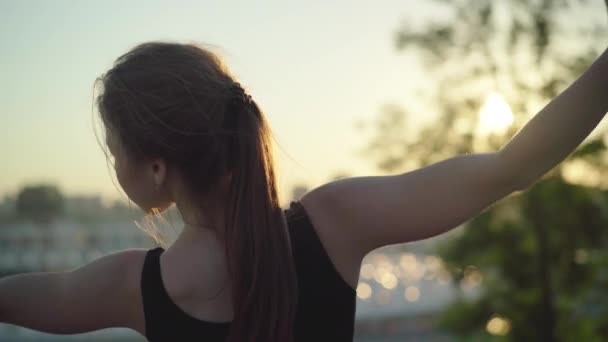 Vista posterior de primer plano de la joven morena entrenando al atardecer con la ciudad urbana en el fondo. Cámara en vivo sigue el movimiento de la esbelta atleta caucásica haciendo ejercicio al aire libre. — Vídeos de Stock