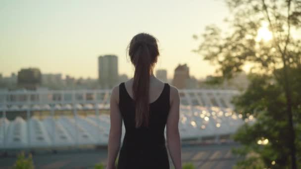 Midden schot van slanke blanke vrouw die bij zonsondergang buiten loopt en naar de camera draait. Portret van een mooie brunette dame poseren in de zomer zonnestralen met stedelijke stad op de achtergrond. — Stockvideo