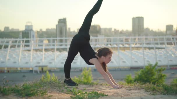 Berdiri split yoga pose wanita ramping fleksibel di luar ruangan sinar matahari. Sisi samping memperlihatkan sorotan luas terhadap olahragawan Kaukasia yang percaya diri yang berolahraga di padang rumput dengan latar belakang kota perkotaan. — Stok Video