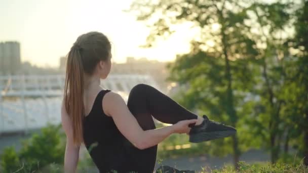 日没時に屋外に座って足を上げる若いスポーツウーマンのバックビュー。都会の夜の街の背景で運動を行うスリム柔軟な女性白人体操. — ストック動画