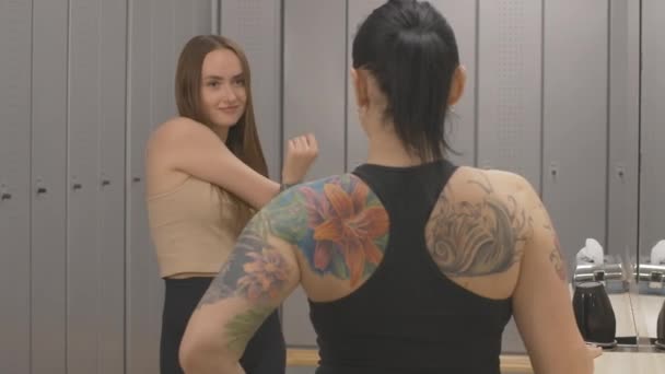 Två tatuerade unga kvinnor chattar i omklädningsrummet innan träning i gym. Porträtt av leende positiva vackra kaukasiska idrottskvinna sträcker ut händerna och lyssnar på vän i omklädningsrummet. — Stockvideo