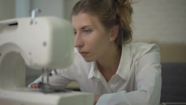 Retrato de cerca de una joven sastre caucásica ajustando la máquina de coser en el taller. Mujer hermosa positiva trabajando con el equipo en el estudio de costura. Concepto de profesiones. — Vídeos de Stock