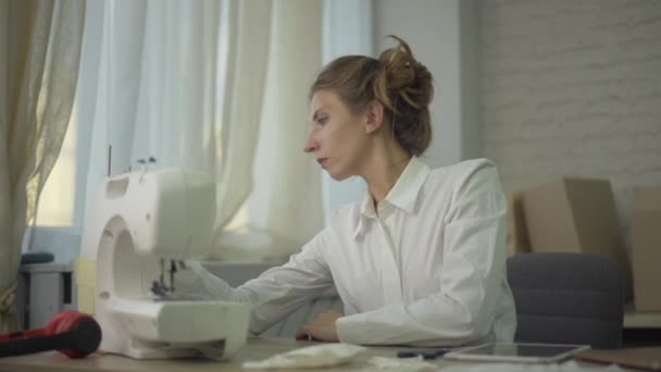 Retrato de mujer caucásica confundida mirando la máquina de coser rota y sosteniendo la cabeza con las manos. Joven sastre morena sentada en el taller y pensando. — Vídeo de stock