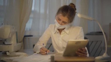 Covid-19 yüz maskeli profesyonel terzi tablet ekrana ve kalemle yazıya bakıyor. Koronavirüs salgını sırasında atölyede kıyafet tasarlayan ciddi bir beyaz kadın..