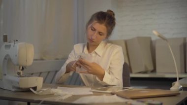 Ciddi bir esmer terzi, Covid-19 yüz maskesini makasla kesiyor, iç çekiyor ve el ele tutuşuyor. Coronavirus virüsü salgını sırasında atölyede çalışan yorgun beyaz kadın portresi.