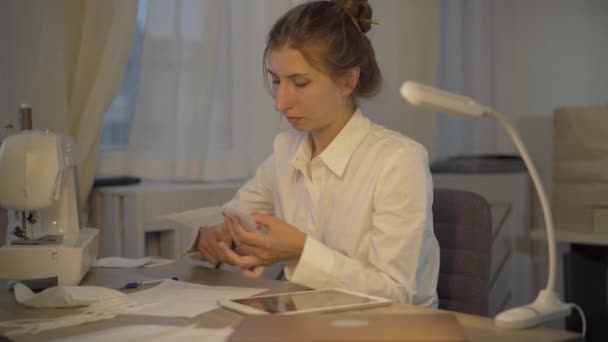 Gericht blanke jonge vrouw snijden Covid gezichtsmasker met een schaar. Zijaanzicht van geconcentreerde naaister of maatwerk ontwerp van coronavirus doek gezicht bedekking. — Stockvideo