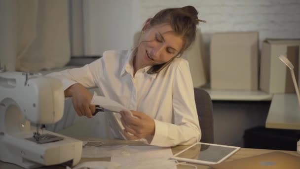 Mujer positiva caucásica cortando mascarilla con tijeras y hablando por teléfono. Retrato de sastre o costurera sentada a la mesa en taller diseñando revestimientos faciales Covid-19. — Vídeo de stock