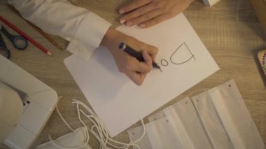 Tanımlanamayan genç bir kadının kağıda bağış yazması. Kadın terzi, yardım için hazırlanmış Covid yüz maskeleriyle masada oturuyor. Gönüllü olma kavramı.