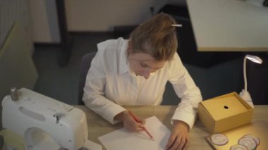 Beyaz kadın, Covid-19 yüz maskesi çiziyor ve kameraya gülümsüyor. Coronavirus sırasında atölyede çalışan kendine güvenen profesyonel terzinin portresi.