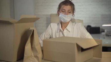 Coronavirus maskeli yorgun bir kadının portresi. Kafasını karton kutulara koyar ve gözlerini kapatır. Covid-19 salgını sırasında kolileri paketleyen yorgun beyaz iş kadınına kamera yaklaşıyor..