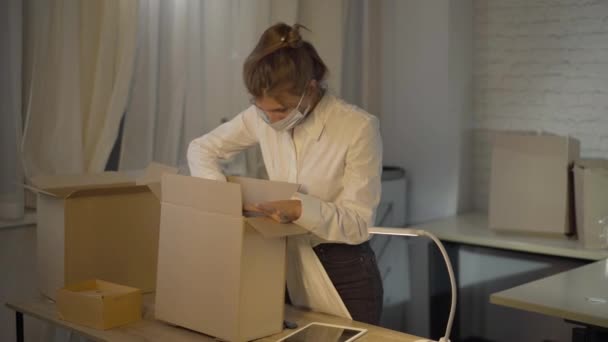 Konzentrierte kaukasische Frau in Coronavirus-Gesichtsmaske Verpackung gut in Atelier zum Verkauf. Konzentrierte Maßschneiderprüfung mittels Tablet und verschließbarem Karton. Covid-19-Kleinunternehmen. — Stockvideo