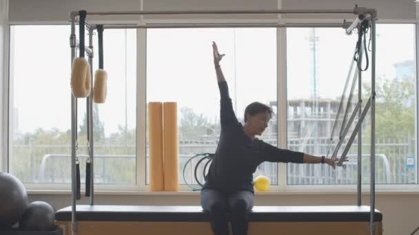 Zelfverzekerde senior Kaukasische sportvrouw zittend op de oefenbank en buigend. Portret van een brunette die traint in de sportschool. Pilates training van serieuze dame in sportclub. — Stockvideo