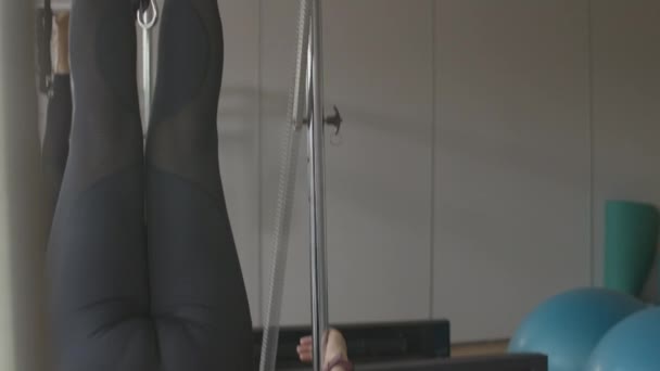 Camera beweegt naar beneden en omhoog langs benen van sportvrouw training op pilates cadillac in de sportschool. Onherkenbare blanke senior vrouw oefenen op sportartikelen binnen. — Stockvideo