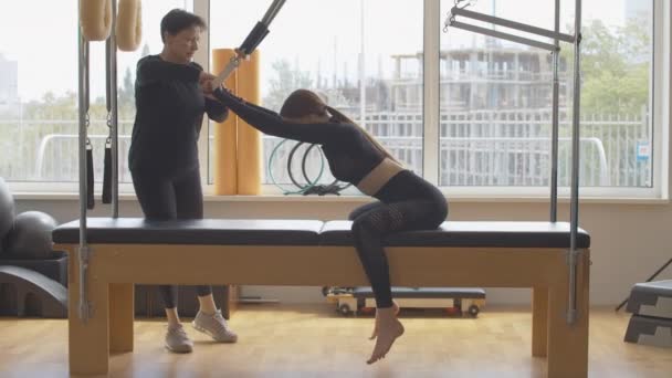 Personlig tränare hjälper unga kaukasiska kvinnan att böja sig på pilates träningsbänk. Brett skott av smal vacker idrottskvinna utbildning i gym med tränare. Träning och idrott. — Stockvideo