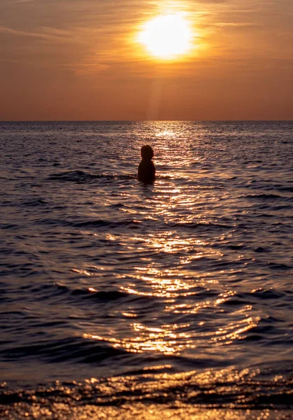 Young woman walks in the waters of Lake Michigan as the sun sets making her a silhouette during this golden hour