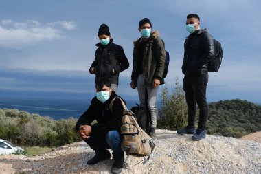 Koruyucu maskeler takan mülteciler ve göçmenler, 4 Mart 2020 'de Yunanistan' ın Mytilene limanına nakledilen askeri otobüsün yanında duruyorlar.