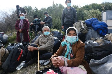 Koruyucu maskeler takan mülteciler ve göçmenler, 4 Mart 2020 'de Yunanistan' ın Mytilene limanına nakledilen askeri otobüsün yanında duruyorlar.