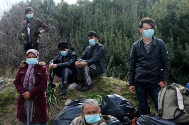 Koruyucu maskeler takan mülteciler ve göçmenler, 4 Mart 2020 'de Yunanistan' ın Mytilene limanına nakledilen askeri otobüsün yanında duruyorlar.