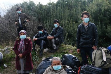 Koruyucu maskeler takan mülteciler ve göçmenler, 4 Mart 2020 'de Yunanistan' ın Mytilene limanına nakledilen askeri otobüsün yanında duruyorlar.