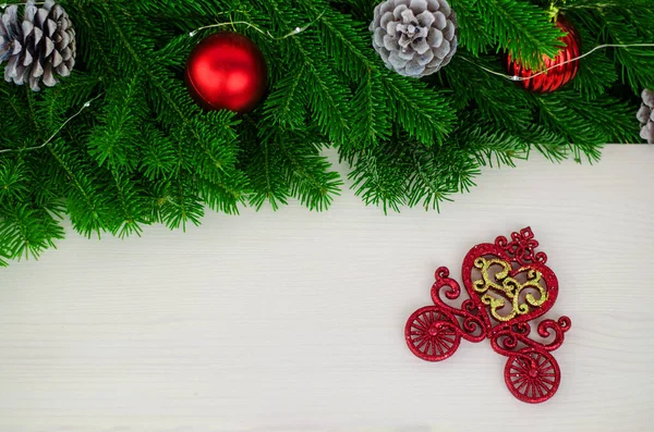 Fir branches are decorated with decorative pine cones and red glass balls and a garland. On a wooden background on the right side is a red sequined decorative carriage.