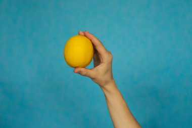 Bir kadının eli mavi arka planda olgun bir limonu tutuyor. Citrus meyvesi. Sağlıklı beslenme.