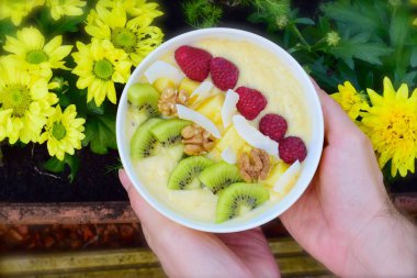 Mango muzlu, ananaslı, ahududu, kivi, ceviz ve hindistan cevizi cipsli kokteyl kasesi tutan kadın elleri.  