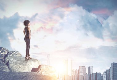 Modern cityscape bulutlu bir gökyüzü ile bir güneşli gün ayakta bir kaya yığını üzerinde seyir emin Afrikalı-Amerikalı kadın. İş ve yaşam tarzı başarı kavramı. Çift tonlu görüntü pozlaması kadar alay