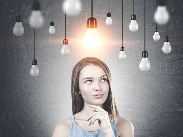 Portrait of a young woman with long fair hair wearing a blue dress and thinking. A blackboard background with many lightbulbs. One is lit.