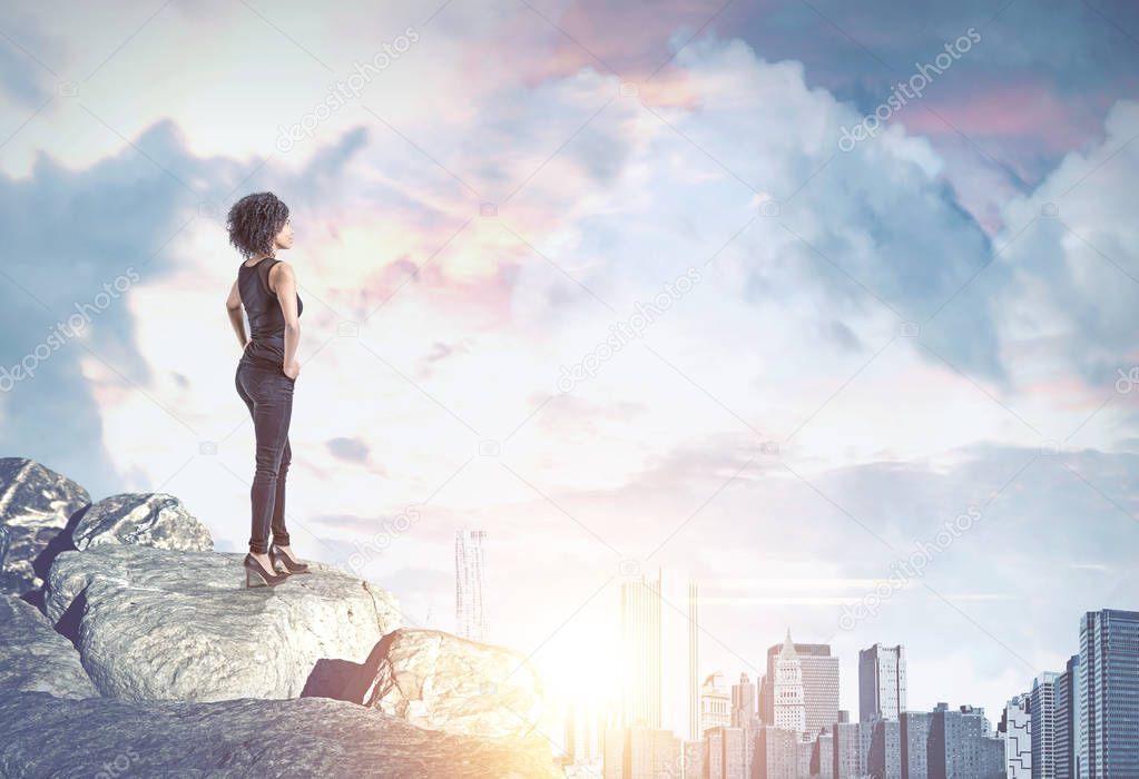 Confident African American woman looking at a modern cityscape with a cloudy sky on a sunny day standing on a pile of rocks. Business and lifestyle success concept. Mock up toned image double exposure