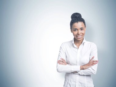 Bir beyaz gömlek ve siyah giyen gülümseyen kendine güvenen genç Afro-Amerikan kadın çapraz kolları ile ayakta ve görüntüleyiciyi seyir pantolon. Gri duvar arka plan sahte