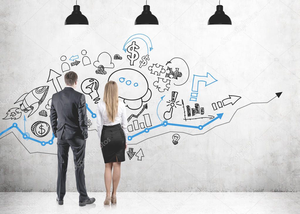 Rear view of young and successful business partners wearing suits looking at a black and blue business profit and financial growth icons on a concrete wall.